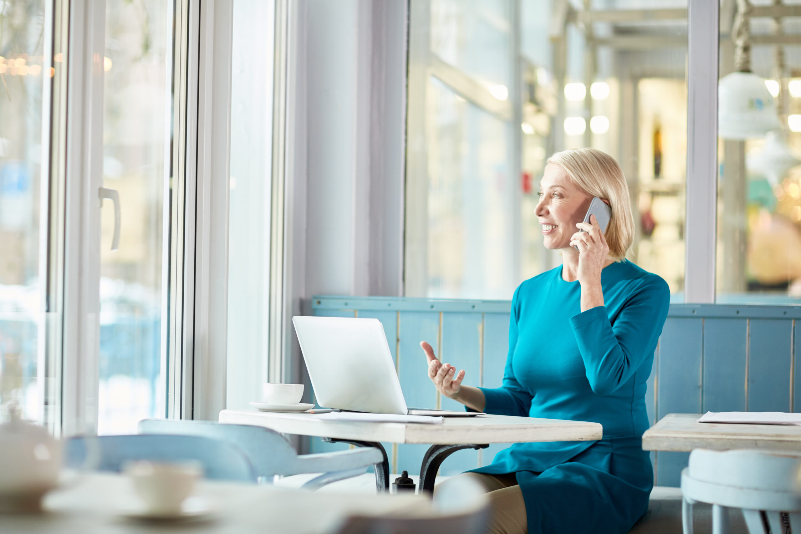 Confident mature businesswoman in smart casualwear sitting by table in cafe in front of laptop and talking to client by smartphone